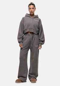 Felpa grigia con zip, maniche oversize e logo frontale. Pantaloni della tuta grigi coordinati con elastico in vita e gamba larga. Scarpe slip-on marroni.