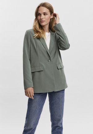 Donna che indossa un blazer verde salvia a un bottone, top bianco e jeans blu, in piedi con una mano che tocca i capelli.