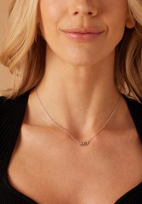 Zilveren ketting met drie onderling verbonden cirkels, hangend aan een delicate ketting tegen een donker geribd topje, wat de huidtint aanvult.
