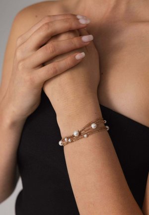 Goldfarbener Kettenarmband mit mehreren Strängen und weißen Perlen, mit einer glatten Textur und einem zarten Design, das sich um das Handgelenk schmiegt.