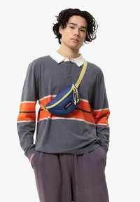 Grå långärmad polo med orange och vita ränder, bärs med en blå midjeväska med gul rem och detaljer. Mjuk tygkvalitet, avslappnad passform.