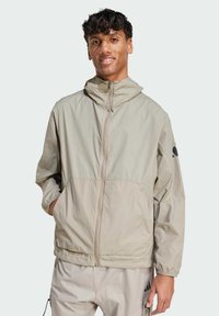 Lightweight beige jacket with a zip closure, elastic cuffs, and hood; features side pockets and a black logo accent on the sleeve.