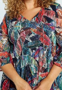 Kihara henkilö, joka on pukeutunut värikkääseen, kuvioituun puseroon, jossa on punaisia, sinisiä, vihreitä ja purppuranvärisiä lehtikuvioita sekä kolmivartiset hihat.