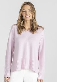 Kobieta z falującymi blond włosami, ubrana w jasnoróżowy sweter w serek i białe spodnie, stoi na tle jednolitego, jasnoszarego tła.