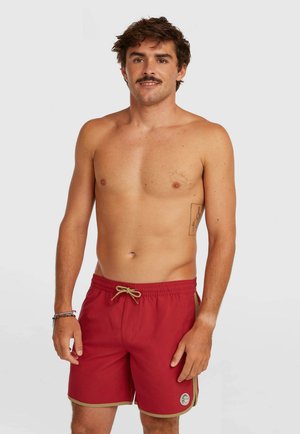 Fitte man met kort bruin haar en snor, gekleed in rode zwembroek met beige rand, staand tegen een effen witte achtergrond.