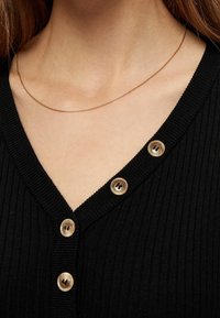 Svart ribbad kofta med V-ringning, prydd med tre ljusbruna knappar fram och ett fint halsband av guldkedja.