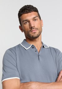 Grijze polo-shirt met een witte bies op de kraag en mouwen. Gemaakt van een textuurstof, voorzien van een knoopsluiting met drie knopen en korte mouwen.