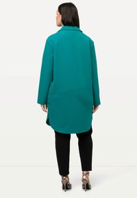Teal kappa med krage, långa ärmar och sidofickor. Kappan har en A-linjeform och når till mitten av låret, i kombination med svarta byxor.