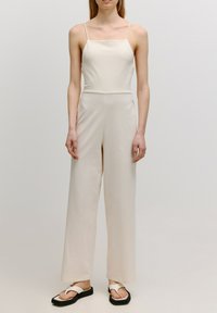 Cremfarbener Jumpsuit mit rechteckigem Ausschnitt, dünnen Trägern und weit geschnittenen Hosenbeinen, aus weichem Stoff. Getragen mit schwarzen Flip-Flops.