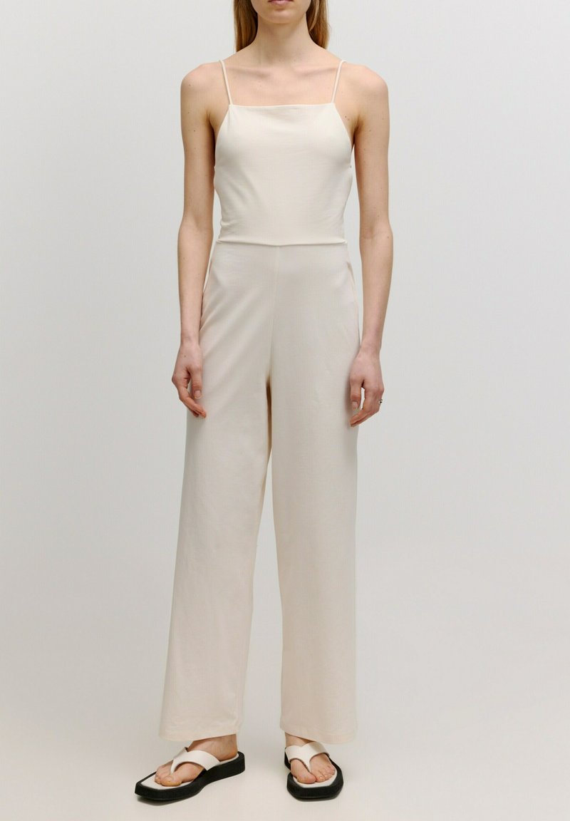 Cremfarbener Jumpsuit mit rechteckigem Ausschnitt, dünnen Trägern und weit geschnittenen Hosenbeinen, aus weichem Stoff. Getragen mit schwarzen Flip-Flops.