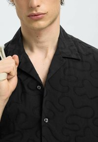 Chemise noire avec un motif texturé et ondulé, col court et deux boutons à l'avant. Le tissu semble léger et doux.