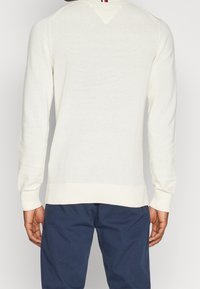 Man in een witte gebreide trui met lange mouwen en donkerblauwe broek, gezien vanaf de achterkant tegen een effen achtergrond.