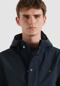 Giovane uomo con capelli corti castani che indossa una giacca blu navy con zip e cappuccio, con un logo ricamato arancione sul petto, su uno sfondo neutro.