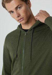 Sudadera con cremallera de color verde oliva, con cordón ajustable, textura suave, bolsillo frontal y diseño ajustado. Tejido liso sin patrones ni logotipos visibles.