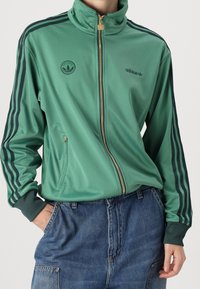 Osoba ubrana w zieloną dresową kurtkę Adidas z czarnymi paskami na rękawach i logo na piersi, w połączeniu z niebieskimi dżinsami.