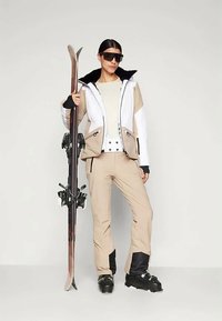 Vrouw in een beige skikleding en zwarte skischoenen, met de ski's rechtop in de hand, draagt grote donkere zonnebril en staat tegen een effen achtergrond.