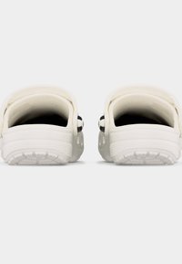 Ζευγάρι λευκών παντόφλων με υφασμένες σόλες και ιμάντες στο πίσω μέρος, τοποθετημένα πάνω σε απλό γκρι ανοιχτό φόντο.