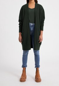 Mörkgrön cardigan med öppen front, ribbade ärmar, tillsammans med en mörkgrön topp, blå jeans och bruna plattformsstövlar.