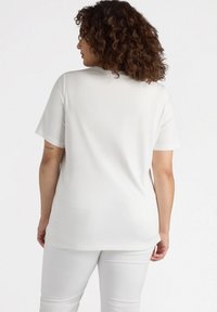 Camiseta blanca de mangas cortas con un corte holgado, hecha de un tejido suave. Presenta un escote redondo y un detalle en la parte trasera, combinada con pantalones blancos.