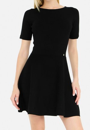 Frau trägt ein kurzärmeliges, figurbetontes schwarzes Kleid mit ausgestelltem Rock und steht vor einem schlichten weißen Hintergrund.