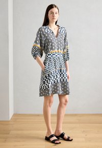 Lyhythihainen mekko, jossa on monivärisiä geometrisiä kuvioita, V-pääntie ja kapeampi vyötärö. Mustat paksupohjaiset sandaalit täydentävät asua.