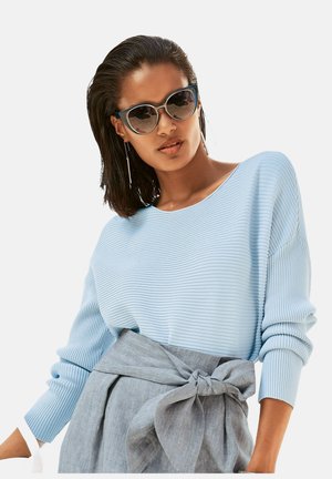 Frau mit kurzen dunklen Haaren trägt Cat-Eye-Sonnenbrille, hellblauen gerippten Pullover und einen grauen Rock mit hoher Taille, der mit großer Schleife gebunden ist.