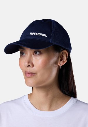 Vrouw met een marineblauwe Rossignol-pet met een achterkant van gaas, kleine creolen en een wit shirt met ronde hals, die lichtjes opzij kijkt.