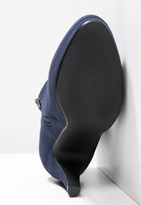 Botín de ante azul marino con tacón alto, detalle de cremallera, suela negra lisa y superficie superior texturizada.