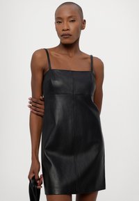Rochie din piele neagră, cu decolteu pătrat și bretele subțiri, având un design ajustat și detalii subtile de cusătură. Textură netedă.