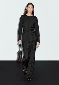 Svart texturerad tvådelad outfit bestående av en figurnära topp med knyt i midjan och vida byxor. Modellen bär en brun handväska och svarta sandaler.