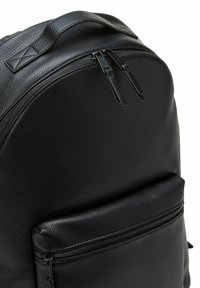 Zaino in pelle nera con texture a grana, forma rotonda, maniglia superiore e tasca frontale con zip. Presenta doppie cerniere nel compartimento principale.