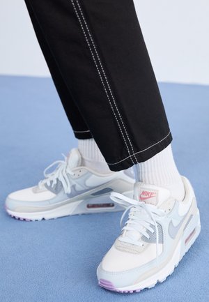 Witte en lichtgrijze Nike Air Max sneakers met roze accenten, gedragen met zwarte broek en witte geribbelde sokken op een blauwe vloer.