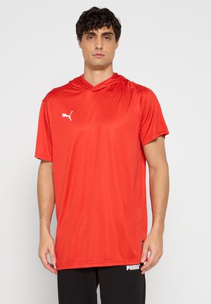Uomo che indossa una maglietta rossa Puma a maniche corte con cappuccio e pantaloni neri Puma in piedi davanti a uno sfondo chiaro e uniforme.