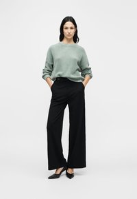 Camisola de malha verde-claro texturizada, gola alta, calças pretas de perna larga, detalhe de botão na cintura, sapatos planos pretos, fundo minimalista.