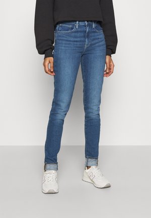 Person wearing blue skinny jeans cuffed at the ankles, white sneakers, and a black long-sleeve top standing on a gray floor.
