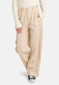 Pantaloni a gamba larga a righe beige e crema con vita alta e pieghe. Indossati con un top bianco a coste a maniche lunghe e sneakers nere.