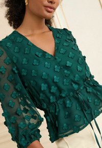 Blusa teal con arricciature floreali, design a V, lunghe maniche trasparenti e vita peplum con dettaglio annodato.