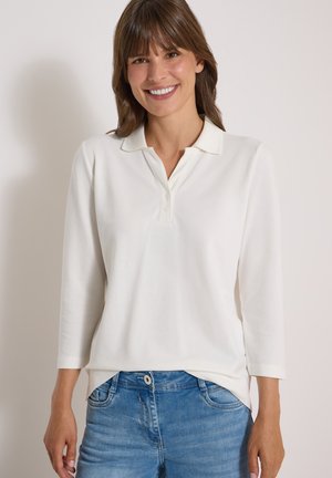 Femme souriante portant un polo blanc à manches longues et un jean bleu sur un fond clair uni.