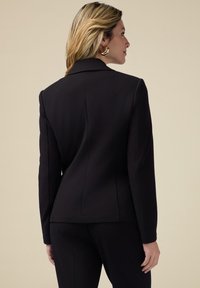 Blazer negru din material croit, cu umeri structurați, deschidere frontală cu un singur nasture și cusături detaliate; purtat cu pantaloni negri asortați.