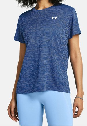 Kobieta ubrana w niebieski sportowy t-shirt z krótkim rękawem z białym logo i jasnoniebieskie dopasowane legginsy, stoi z ramionami swobodnie opuszczonymi wzdłuż ciała.
