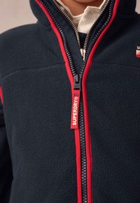 Tamsiai mėlynas vilnonis švarkelis su raudonomis detalėmis, užtrauktuku ir firminiu užtrauktuko traukikliu. Turi aukštą apykaklę ir minkštą tekstūrą.