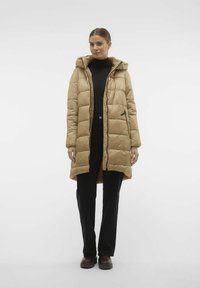 Beige gewatteerde jas met capuchon, voorzien van een rits aan de voorkant en zwarte accenten. Lange mouwen en zijzakken. Draag het over een zwarte coltrui en broek.