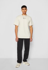 Beige cotton t-shirt with short sleeves and a black handwritten logo on the chest, paired with black pants and cream sneakers.