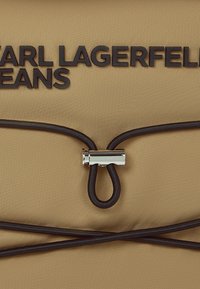 Beige tygpåse med svart text "CARL LAGERFELD JEANS", silverfärgad hårdvara och svarta snöraccenter för stängning. Slät textur.