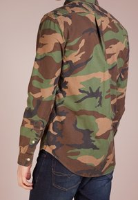 Camisa de manga larga con estampado de camuflaje en verde, marrón y negro. Cuenta con un cuello, puños con botón y dobladillo curvado. Fabricada en suave tejido de algodón.
