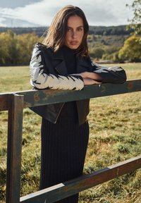 Zwarte leren jas met unieke patroonmouwen, gedragen over een geribbelde zwarte jurk. De achtergrond toont een groen veld en bomen.