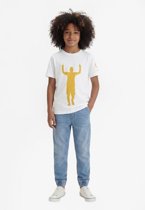 Enfant aux cheveux bouclés portant un t-shirt blanc avec une silhouette jaune et un jean bleu clair, debout avec une main dans la poche.