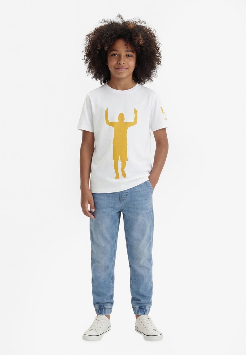 Enfant aux cheveux bouclés portant un t-shirt blanc avec une silhouette jaune et un jean bleu clair, debout avec une main dans la poche.