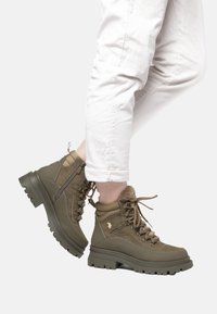 Olivgröna ankelstövlar i texturerad mocka och läder, med vadderad krage, snördesign och en robust svart gummisula med djup mönstring.