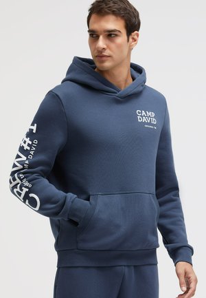 Mann trägt einen dunkelblauen Hoodie mit dem Schriftzug „CAMP DAVID“ auf Brust und linkem Ärmel, Hände in der Fronttasche, schaut zur Seite.